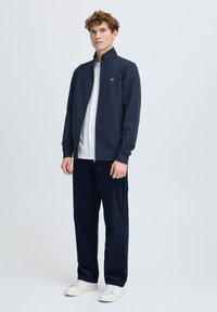 Marineblå zip-up jakke med høy krage, laget av bomullsblanding, med en liten logo på brystet, sammen med en hvit t-skjorte og mørke bukser.