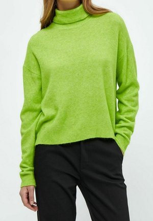 Femme portant un pull vert citron vif à manches longues avec col montant et pantalon noir, debout avec une main dans la poche.