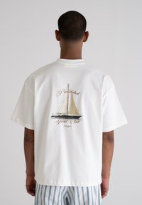 White cotton t-shirt featuring a sailboat graphic on the back, with "Prohibited Yacht Club" text above and "Grand Resort" below in a classic font.