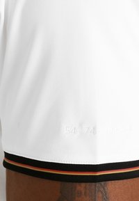 Vit kortärmad skjorta med en texturerad tyg, med svart, röd och gul kant längst ner och präglade siffror på sidan.