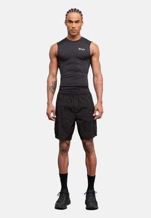 Joven con cabello trenzado que lleva una camiseta negra sin mangas ajustada, pantalones cortos cargo negros, calcetines negros y zapatillas negras, de pie mirando hacia adelante.