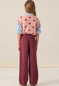Gilet en laine rose avec des motifs en forme de cœur rouge sur une chemise à rayures bleu, associé à un pantalon large bordeaux. Superposition claire et décontractée évidente.
