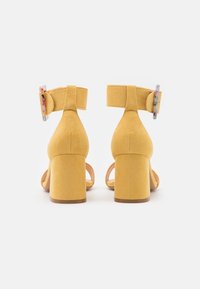 Keltaiset mokka sandaalit kannalla, jossa on paksu korko, nilkkavarsi ja koristeellinen solki, jossa on monivärisiä yksityiskohtia.