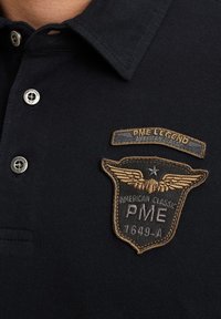 Mornarska polo majica s ovratnikom, slična srebrenim gumbima i istaknutom izvezenom oznakom s krilima te tekstom u zlatnoj i bijeloj boji.