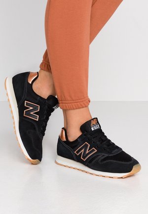 Osoba nosząca czarno-różowo-złote sneakersy New Balance 373 z białymi podeszwami i brązowym bieżnikiem, w połączeniu z pomarańczowymi spodniami dresowymi.