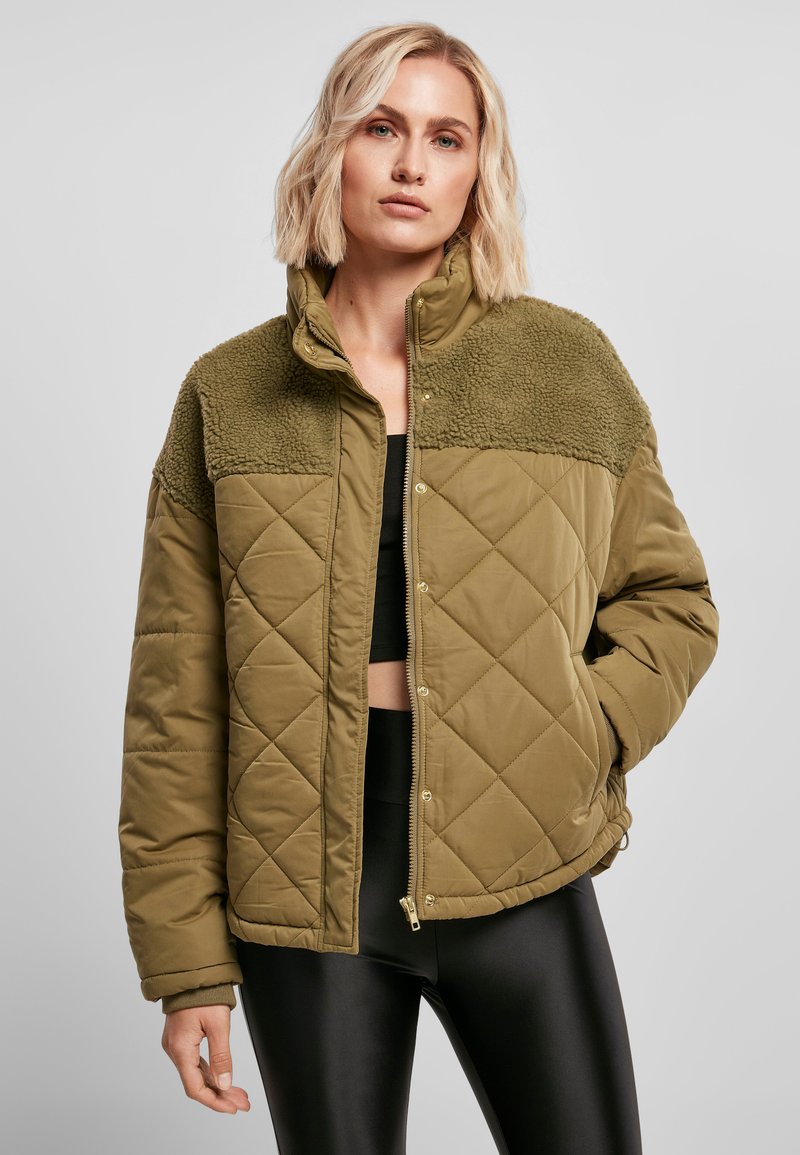 Olivengrønn quiltet jakke med pelskrage, med glidelås og trykknapper, kombinert med svarte leggings.