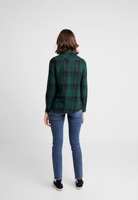 Camicia verde a quadri con maniche lunghe, polsini abbottonati e colletto, abbinata a jeans slim blu e sneakers nere.