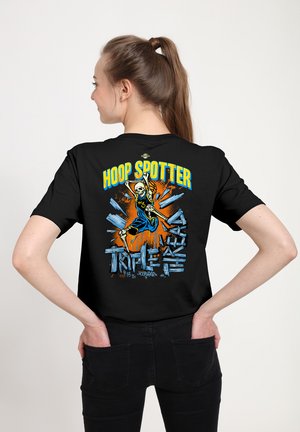 Osoba o brązowych włosach związanych w kucyk, ubrana w czarne spodnie i czarny T-shirt z nadrukiem szkieletowego koszykarza oraz napisami "Hoop Spotter" i "Triple Threat".