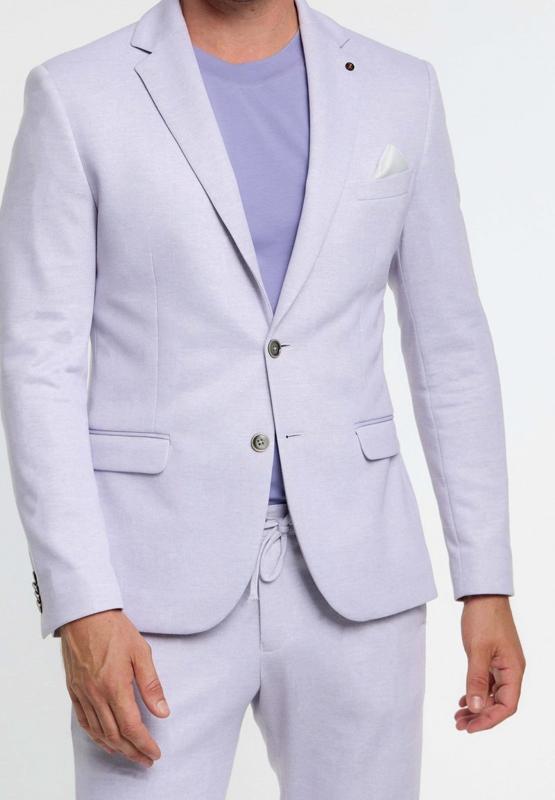 Homme portant un blazer lavande clair avec un mouchoir de poche, un pantalon à cordon assorti et un t-shirt lavande à col rond.