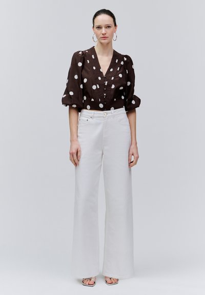 Naine, kes kannab tumepruuni pluusi valgete täppidega, valgeid kõrge vöökohaga laia säärega pükse, rõngakujulisi kõrvarõngaid ja avatud varbaga sandaale, seisab.