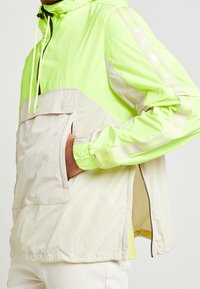 Lekka kurtka z kapturem, z żywym neonowo-zielonym górnym fasonem i beżowym dolnym, z odblaskowymi akcentami oraz przednią kieszenią.