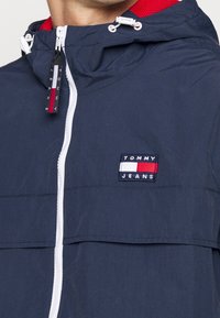 Marinblå lättviktsjacka med vit dragkedja och snoddar, med en röd och vit fodrad huva. Broderad "TOMMY JEANS"-logotyp på bröstet.