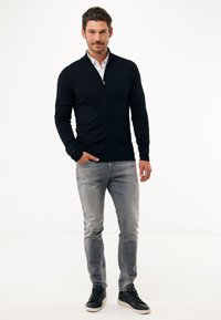 Černá zipová mikina s vysokým límcem, nošená přes bílou košili, spárovaná s světle šedými džíny a černými teniskami. Jednoduchý design a přiléhavý střih.