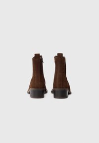 Bottes de cheville en suede marron avec une texture lisse, fermeture éclair et un petit talon carré. Bout rond et design minimaliste.