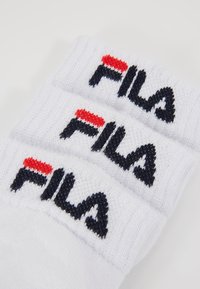 Tri parova bijelih pamučnih sportskih čarapa s pletenom teksturom i Fila logotipom u crnoj i crvenoj boji na rubu.