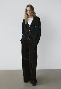 Zwarte gebreide cardigan met twee borstzakken, gouden knopen; gecombineerd met wijd uitgesneden donkerbruine ribfluwelen broek, gladde textuur, en een eenvoudig wit topje.