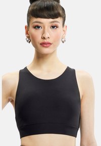 Young woman with dark hair in a bun wearing hoop earrings and a sleeveless black crop top, facing forward against a plain white background.