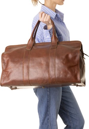 Persona con camicia a righe blu e jeans che porta a mano una grande borsa da viaggio marrone in pelle.