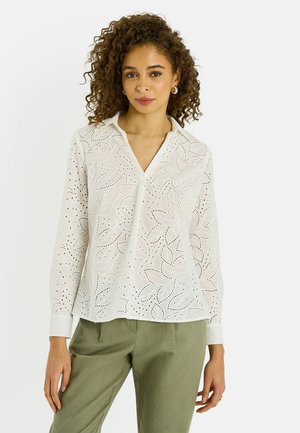 Femme aux cheveux bouclés portant des boucles d'oreilles dorées, un chemisier à manches longues blanc avec des ajourés floraux, et un pantalon vert olive.