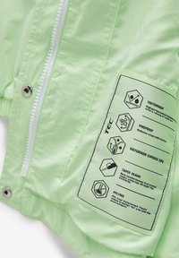 Hellgrüne, wasserdichte Jacke mit einem weißen Reißverschluss, ausgestattet mit einem Etikett, das die Leistungsmerkmale und Pflegehinweise beschreibt. Strukturierter Stoff.