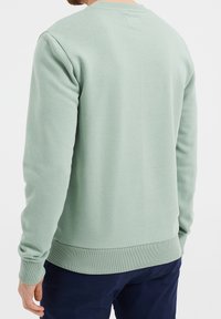 Muž v světlezeleném dlouhém svršku a tmavě modrých kalhotách, zobrazený zezadu na bílém pozadí.