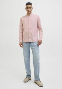 Ljust rosa skjorta med ståkrage, i kombination med ljusblå jeans och beige slip-on skor, mot en enfärgad bakgrund.