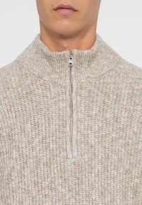 Mies, joka pukeutuu vaaleanbeessiin ja harmaan kirjavavilla neulepaidalle, jossa on korkea kaulus ja puolivetoketju hopeavärillä, paljastaen kaulan ja alanuoren kasvot.