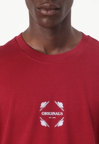 Red cotton t-shirt featuring a crew neck, short sleeves, and a white print with the word "ORIGINALS" and decorative leaf designs.