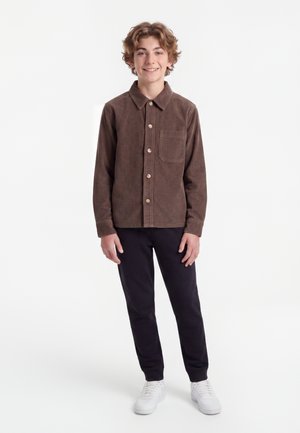 Smilende teenage dreng med krøllet hår iført en brun skjorte med knapper, sorte bukser og hvide sneakers, står foran en hvid baggrund.