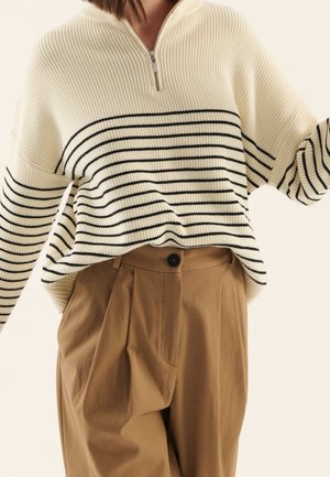 Kvinde iført en cremefarvet ribstrikket sweater med sorte vandrette striber og tanfarvede højtaljede plisserede bukser med en sort knap.