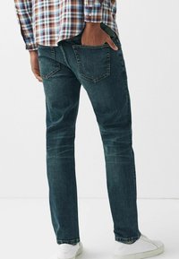 Tummansiniset denimfarkut kapealla istuvuudella, joissa on perinteinen viiden taskun malli ja hienovarainen haalistuma, yhdistetty valkoisiin tennareihin.