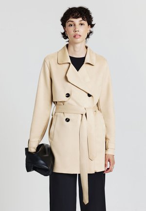 Donna con capelli corti e ricci, indossa un trench coat beige con cintura e pantaloni neri, tiene in mano sinistra una borsa clutch nera piegata.