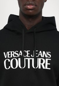 Μαύρη φούτερ μπλούζα με τη φράση "Versace Jeans Couture" εκτυπωμένη με μεγάλα λευκά γράμματα στο στήθος, φορε�μένη από ένα άτομο με σκούρο δέρμα.