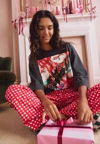 Black graphic t-shirt featuring a colorful holiday design, paired with red and white checkered pajama pants. Posing with a pink gift box.