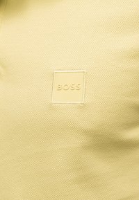 Tissu jaune texturé avec un petit patch carré cousu au centre affichant le mot « BOSS » en jaune assorti.