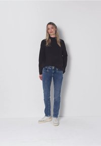 Černý svetr, modré džíny a bílé tenisky. Svetr má dlouhé rukávy a džíny jsou přiléhavé se středním vypráním.