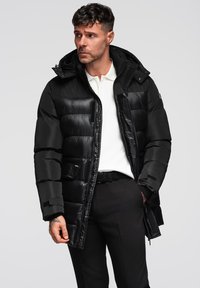 Giacca a vento nera con cappuccio, caratterizzata da una texture trapuntata, chiusura a zip e tasche. Abbinata a una polo bianca e pantaloni neri.