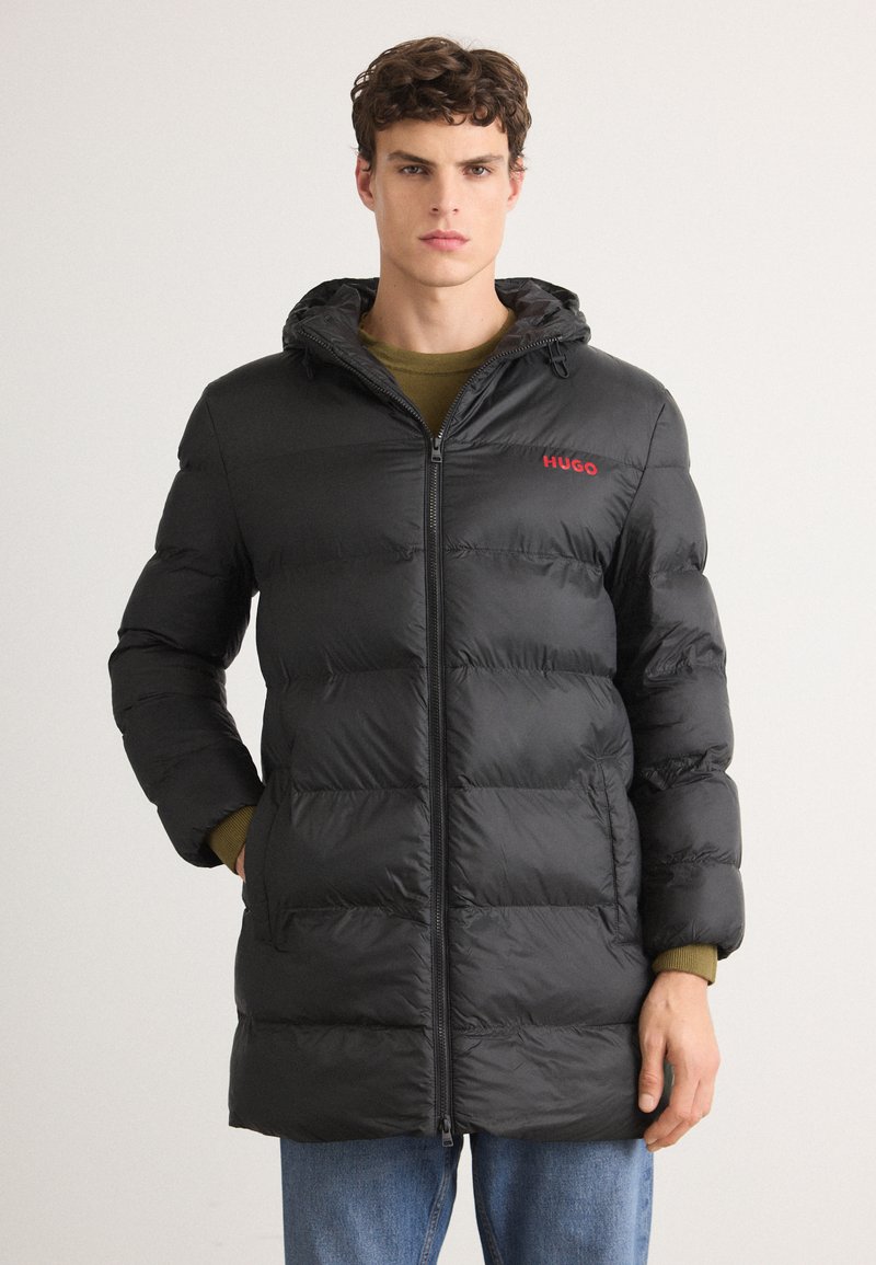 Chaqueta de plumas negra con cremallera frontal, capucha y logo rojo. Cuenta con secciones acolchadas y un corte ligeramente alargado para mayor calidez.
