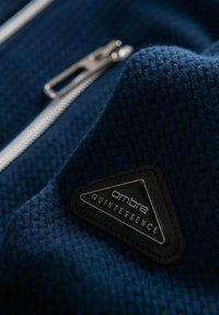 Marinblått strukturerat tyg med ett stickat mönster, som har en silverdragkedja och en svart triangulär varumärkesetikett med texten "ombre QUINTESSENCE."