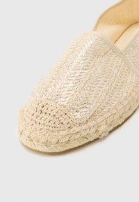 Beige vevd espadrille med metalliske detaljer, rund tå og en flettet jute-såle. Har en heklet detalj ved tåhetten.