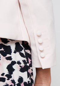 Světle růžový blazer s hladkou texturou, který má čtyři kulaté knoflíky na manžetě. V kombinaci s sukni s multicolor květinovým vzorem.