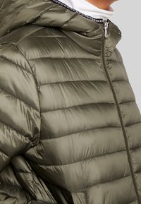 Olivgrüne Steppjacke mit glänzender Oberfläche, horizontal gestepptem Muster und durchgehendem Reißverschluss. Mit Kapuze und kontrastierendem Besatz am Hals.