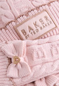Lyserød strikket hue med kabelstrikkemønster, der har en tekstureret sløjfe med en guldfarvet hjerteknap og et brandmærke syet med guldfarvet tråd.