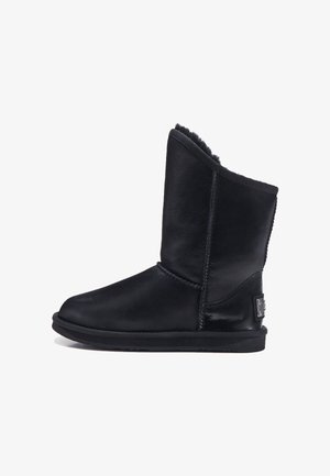 Bota de cuero negro hasta el tobillo con forro suave, parte superior curvada y suela de goma plana. Detalle de costura notable y acento metálico con marca en el lateral.