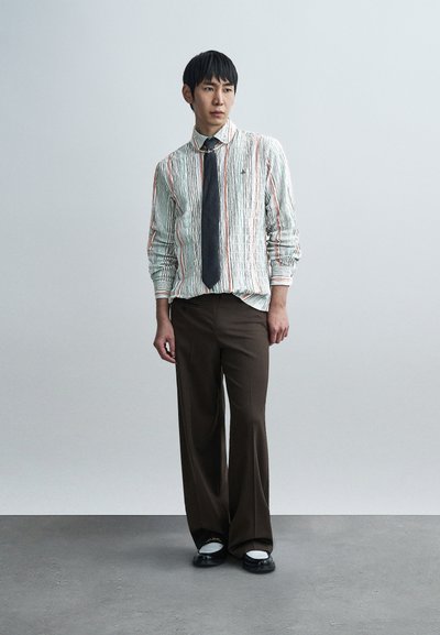 Άνδρας που φοράει ρυτιδωμένο ριγέ πουκάμισο με μαύρη γραβάτα, καφέ φαρδιά παντελόνια και ασπρόμαυρα παπούτσια στέκεται μπροστά σε ένα απλό φόντο.