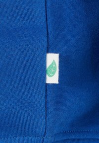 Blauer Strickstoff mit einem kleinen weißen Etikett, das ein grünes Blattsymbol zeigt und in eine Naht eingenäht ist.