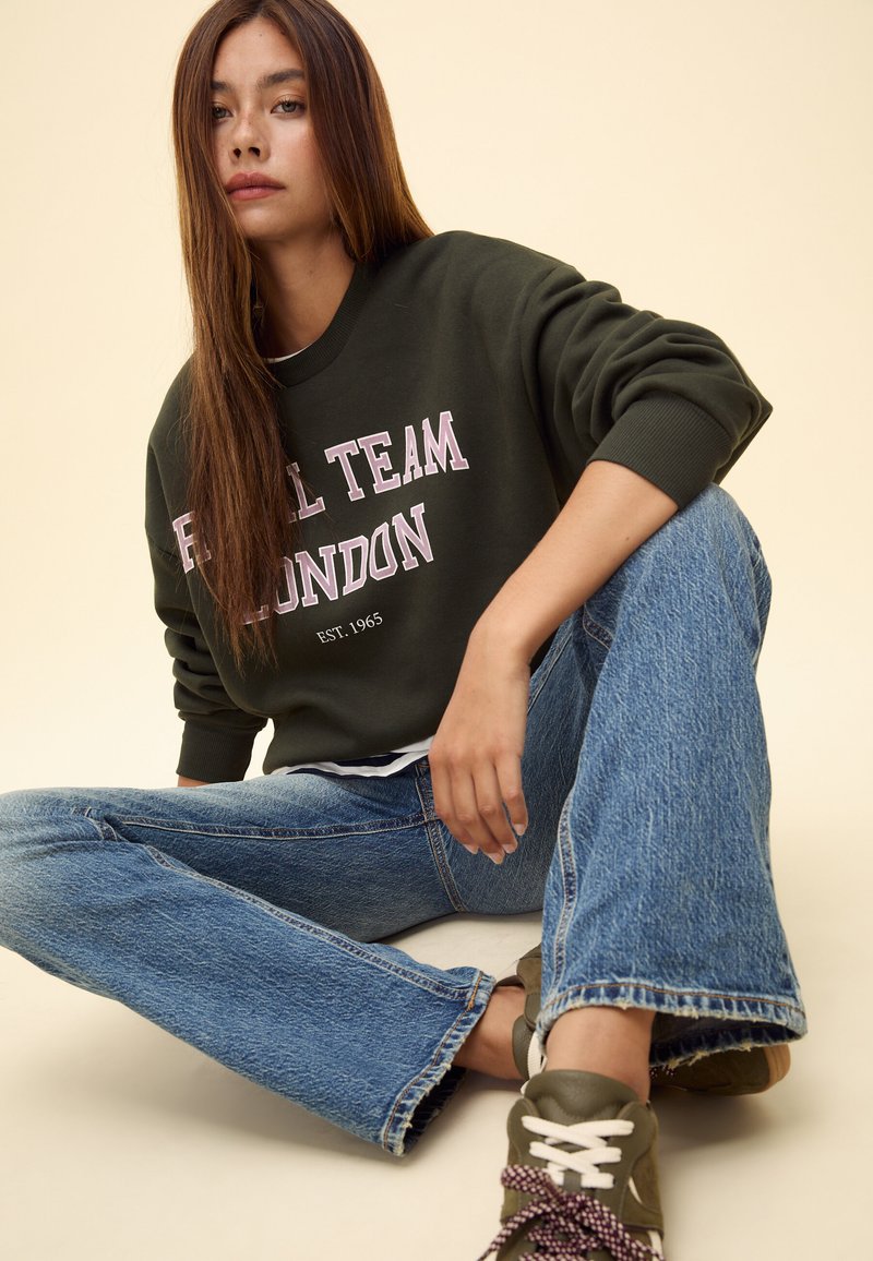 Giovane donna con lunghi capelli castani che indossa una felpa scura con la scritta "Royal Team London" e jeans blu, seduta a gambe incrociate.