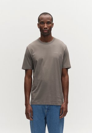 Mees, kes kannab lihtsat pruuni lühikeste varrukatega T-särki, millel on "GABBA" logo, ja siniseid teksaseid, seisab ühevärvilise valge tausta ees.