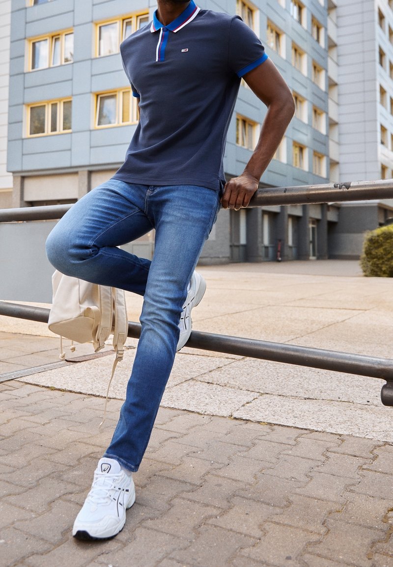 Polo bleu marine avec col contrastant, jean denim bleu et baskets blanches. La personne s'appuie sur une rambarde, tenant un sac à dos crème.