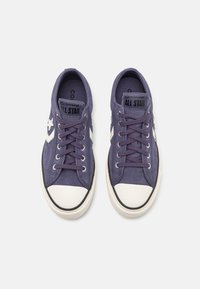 Fioletowe trampki Converse All Star w niskim wzorze z białymi gumowymi czubkami, białym logo gwiazdy po bokach i fioletowymi sznurówkami na białym tle.